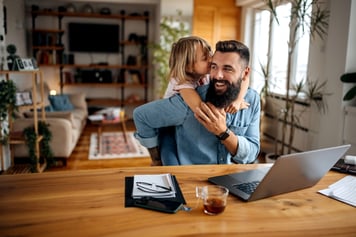 Daughter hugging father in living room while father sits at table working on laptop