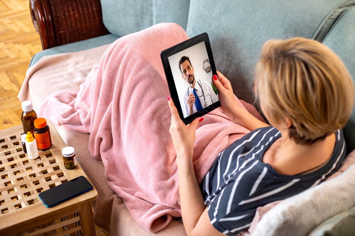 Woman laying on couch under pink blanket using tablet for video telemedicine call with her mle doctor