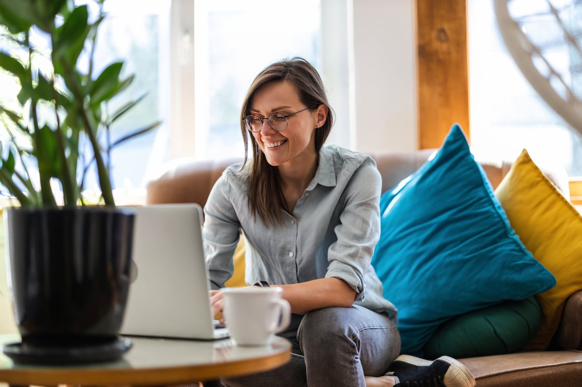 Woman with glasses working on laptop while sitting on brown sofa