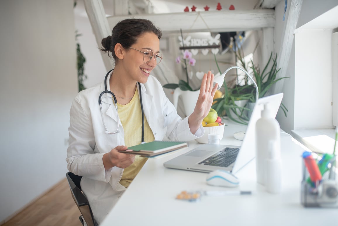 Doctor in labcoat waving and speaking to patient through computer telemedicine call.